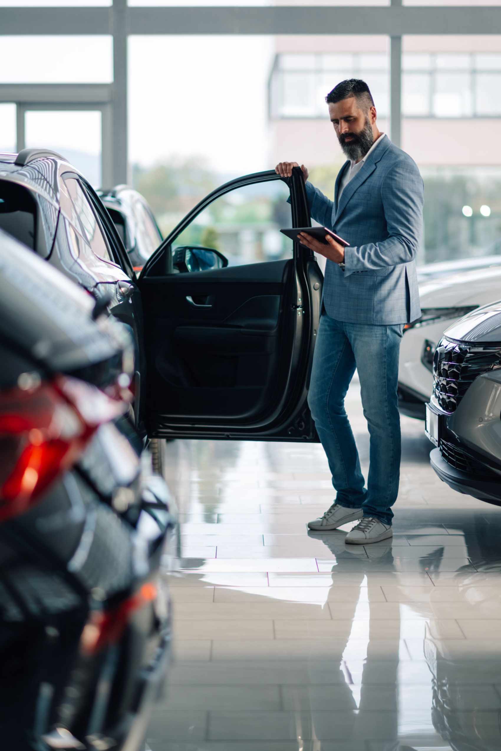 Autoverkäufer mit digitalem Tablet, der dem Kunden im Ausstellungsraum des Händlers die Merkmale des Neuwagens zeigt
